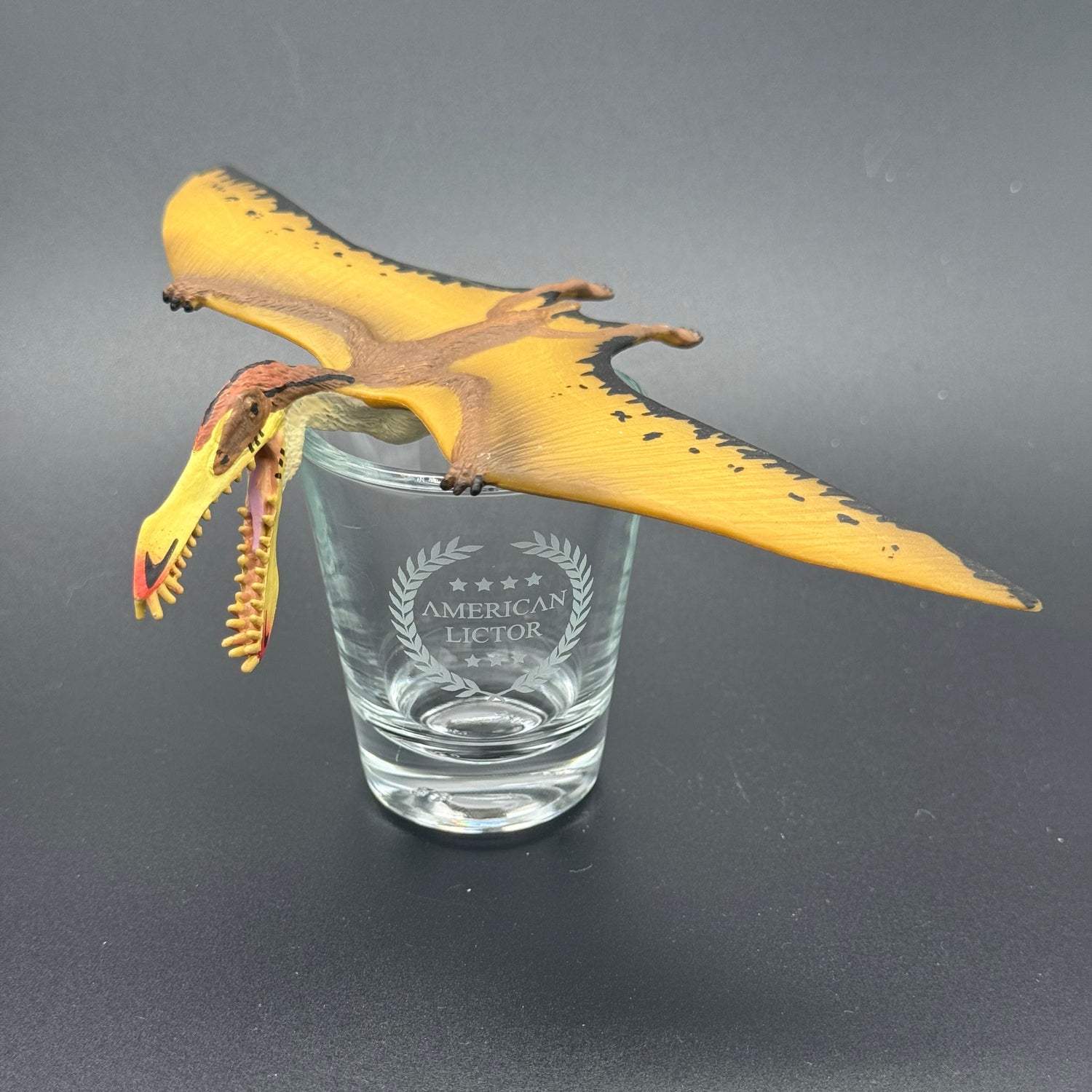A Shot glass with the American Lictor Logo posed with a dinosaur