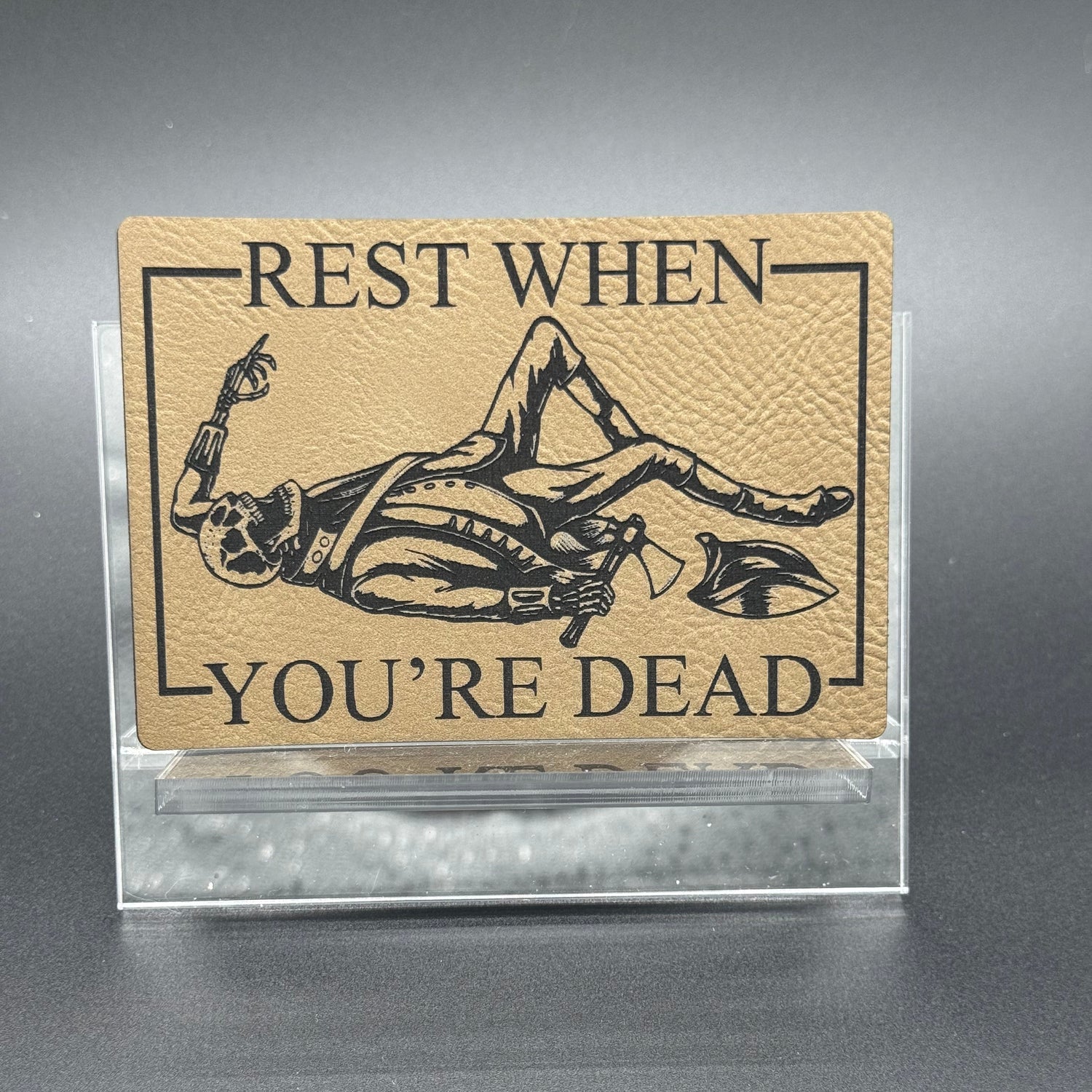A leatherette patch in beige picturing a skeleton wearing colonial soldier clothing with a tomahawk and the text 'Rest when you're dead