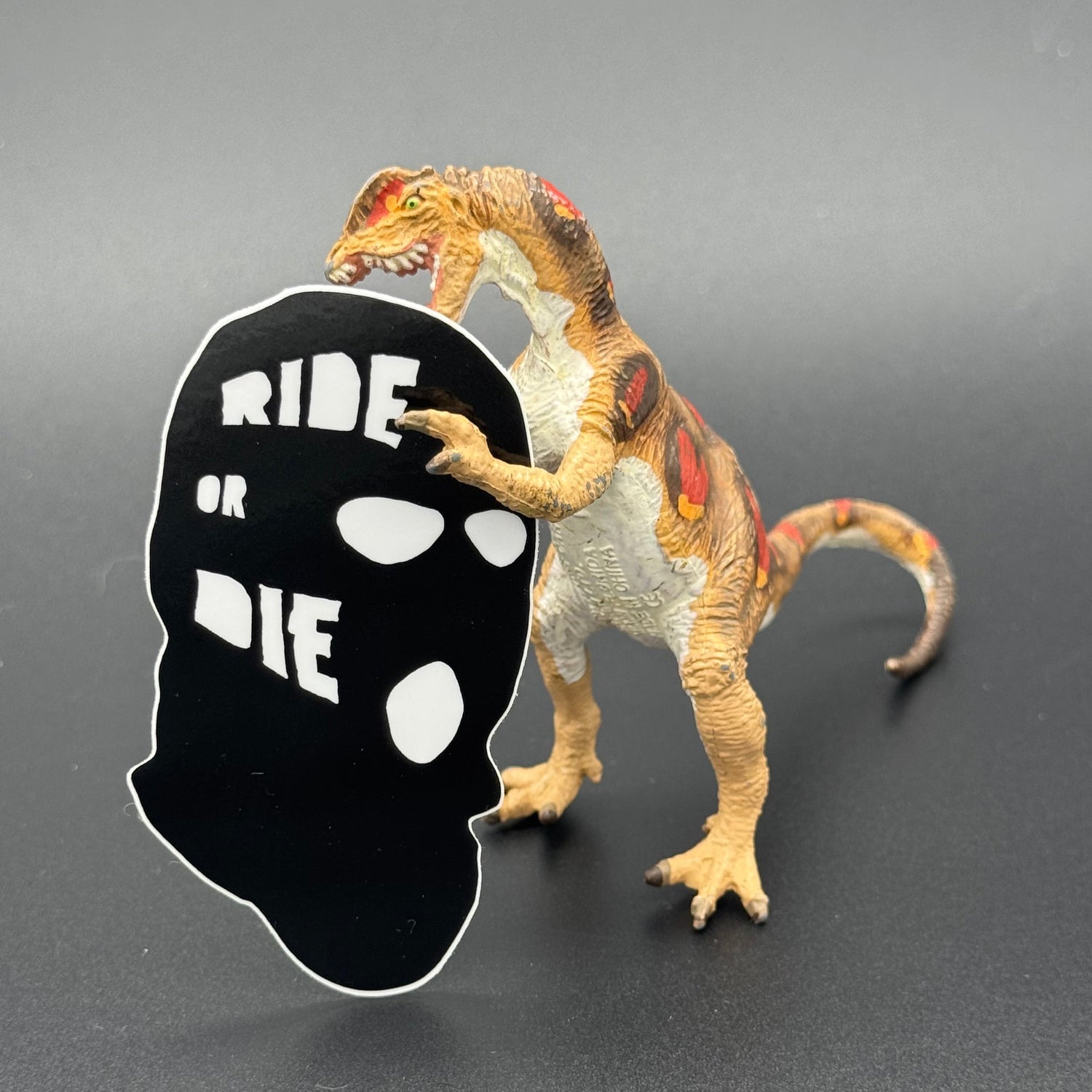 Sticker of a black ski mask with the text 'Ride or Die' posing with a dinosaur