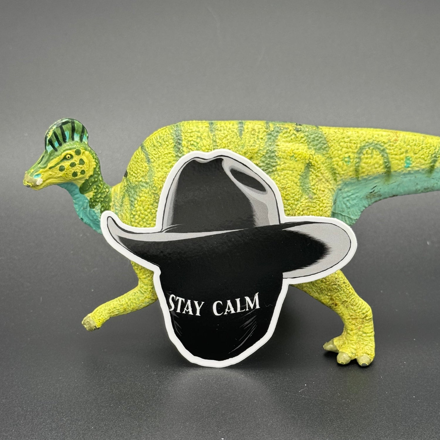Sticker of a masked man with a black cowboy hat and the text 'Stay Calm' posing with a dinosaur