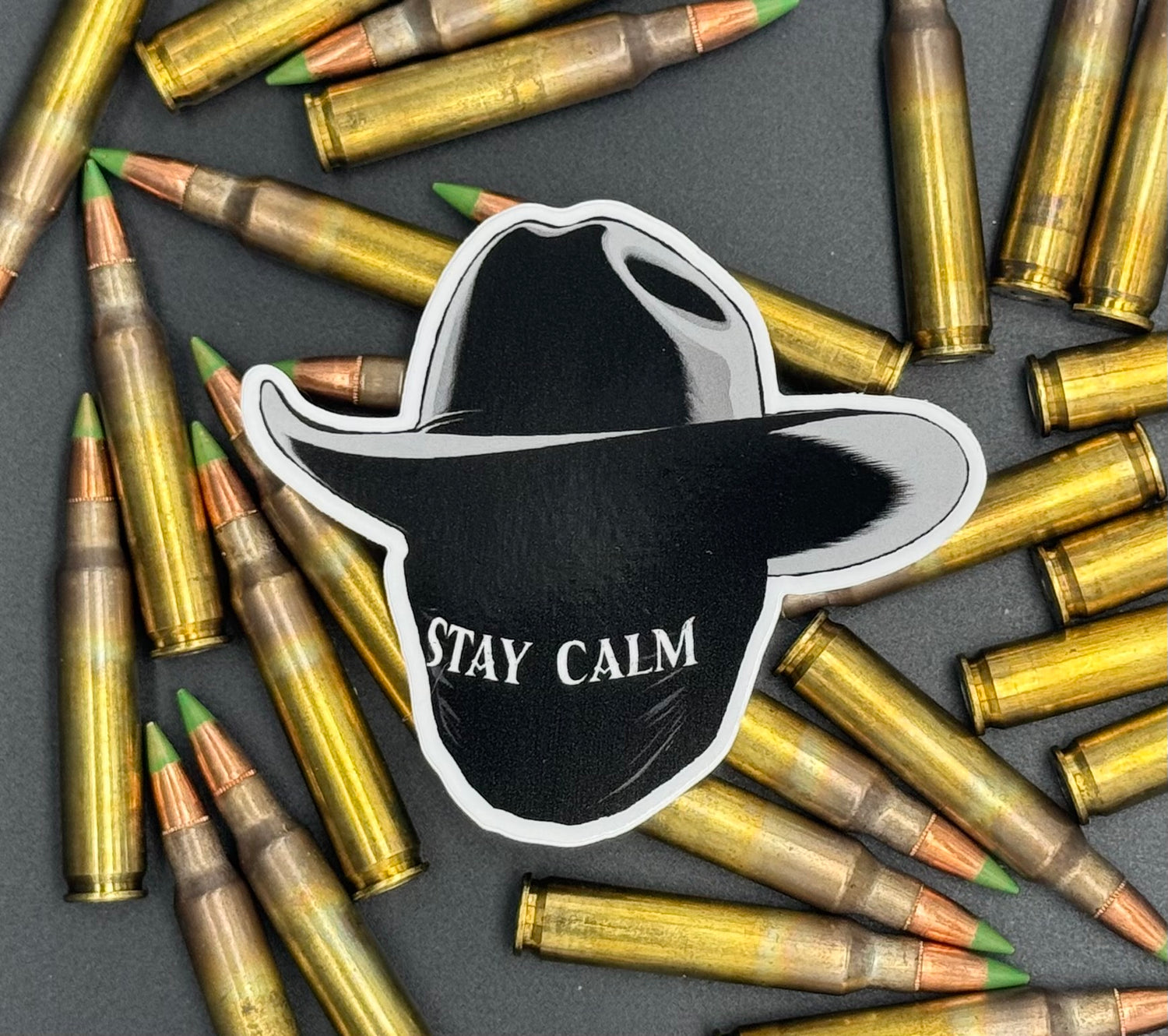 Sticker of a masked man with a black cowboy hat and the text 'Stay Calm' on a bed of ammo