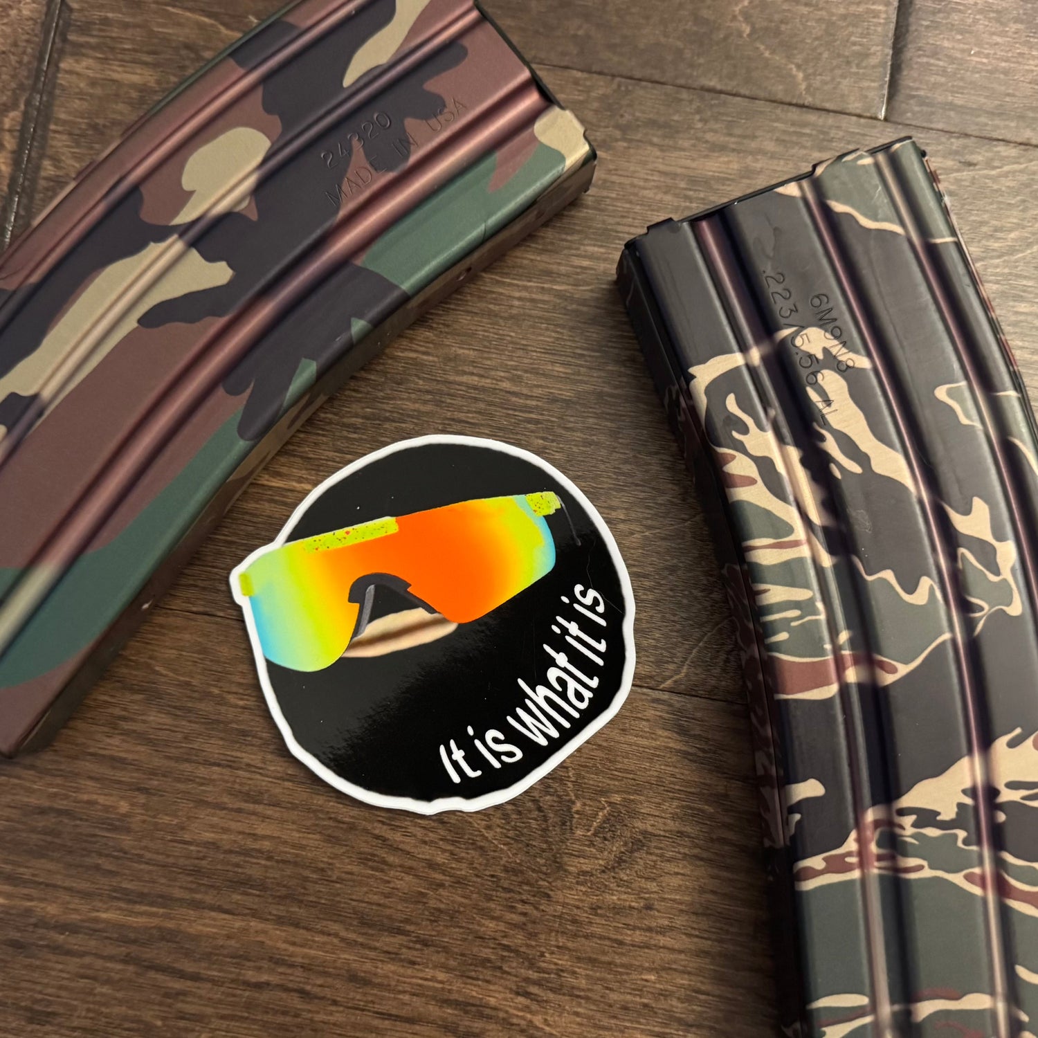 Camouflage magazine on a wooden surface with a sticker featuring sunglasses and text 'It is what it is'