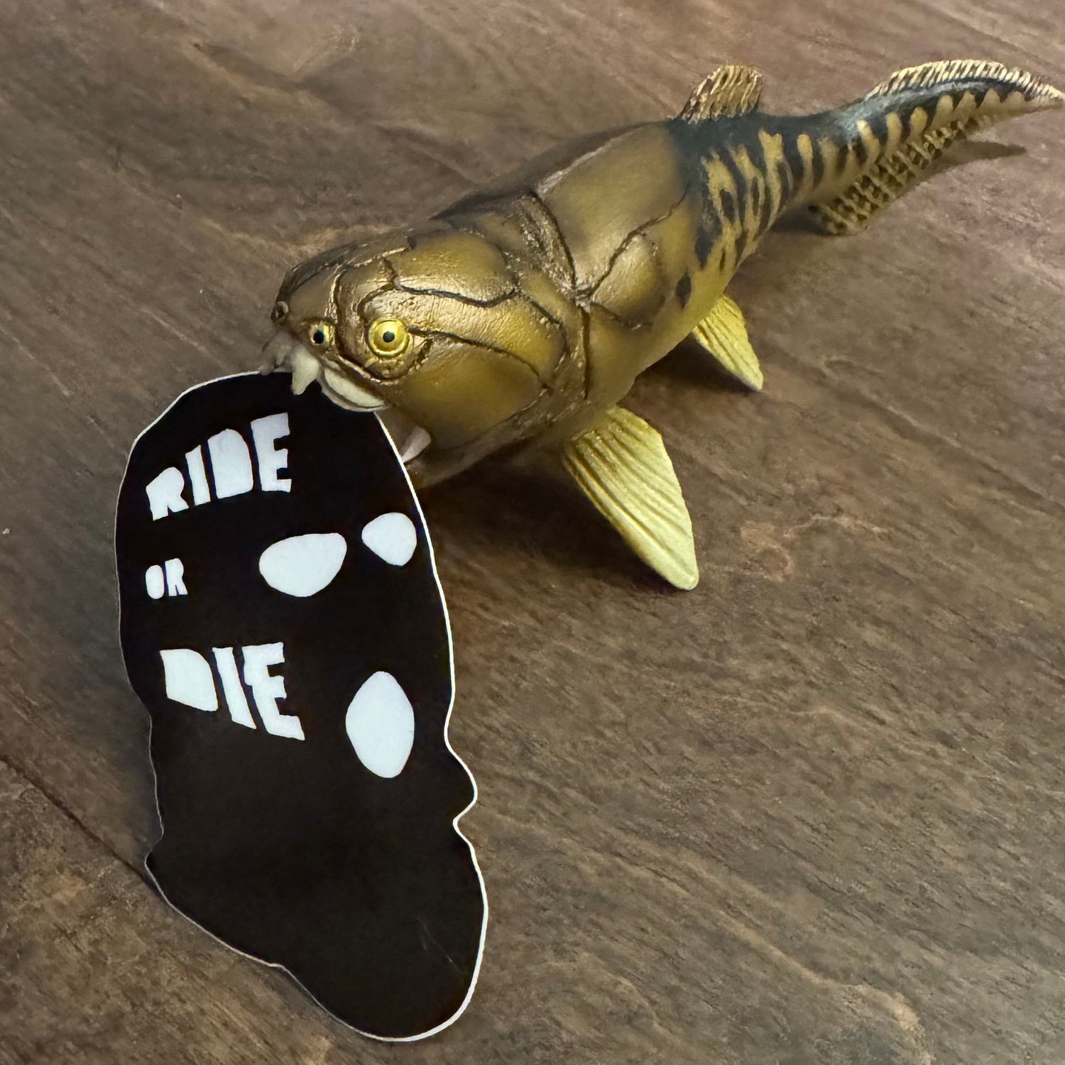 Toy dinosaur with a 'Ride or Die' sticker on a wooden surface