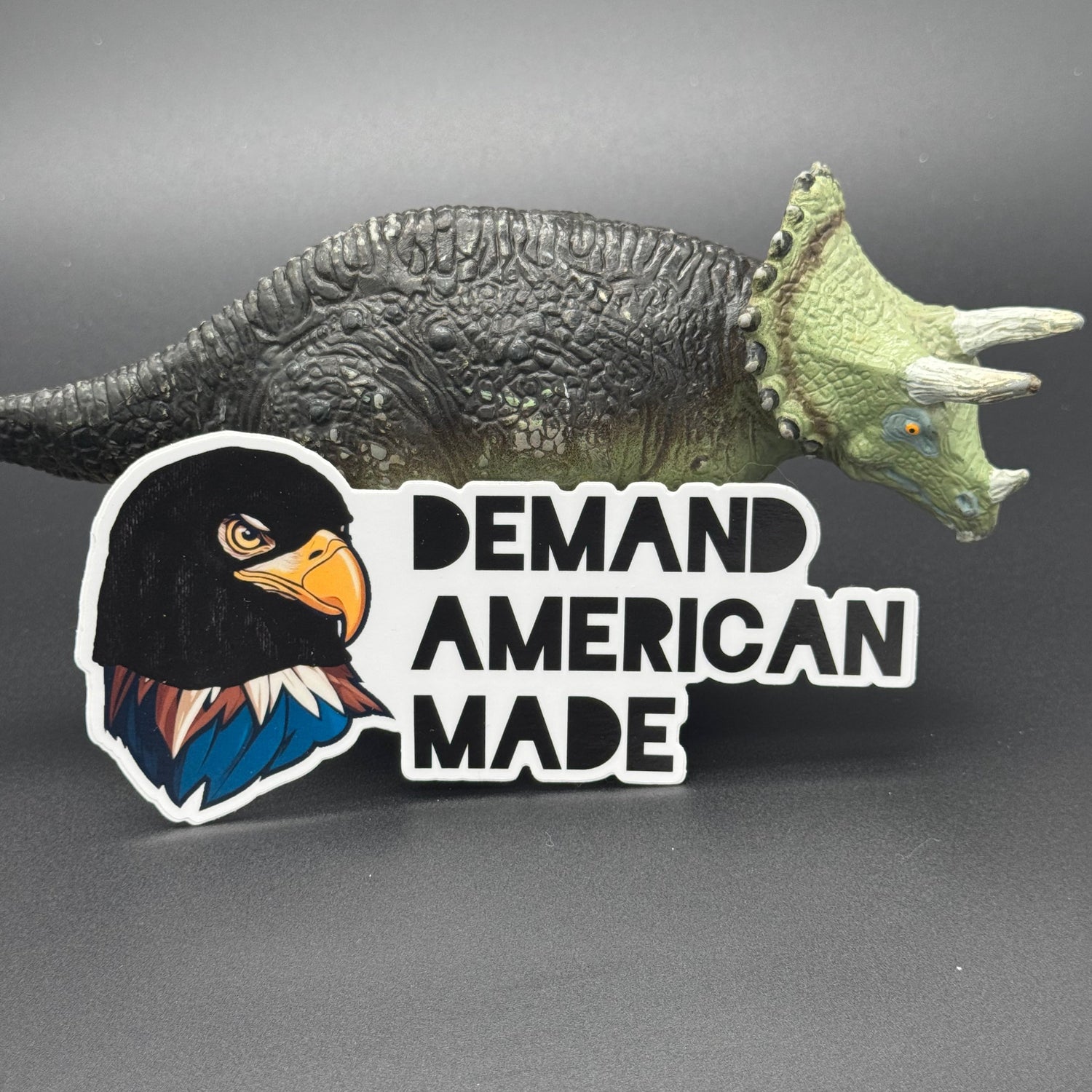 Sticker of an Eagle wearing a ski mask with the text 'Demand American Made' posing with a dinosaur