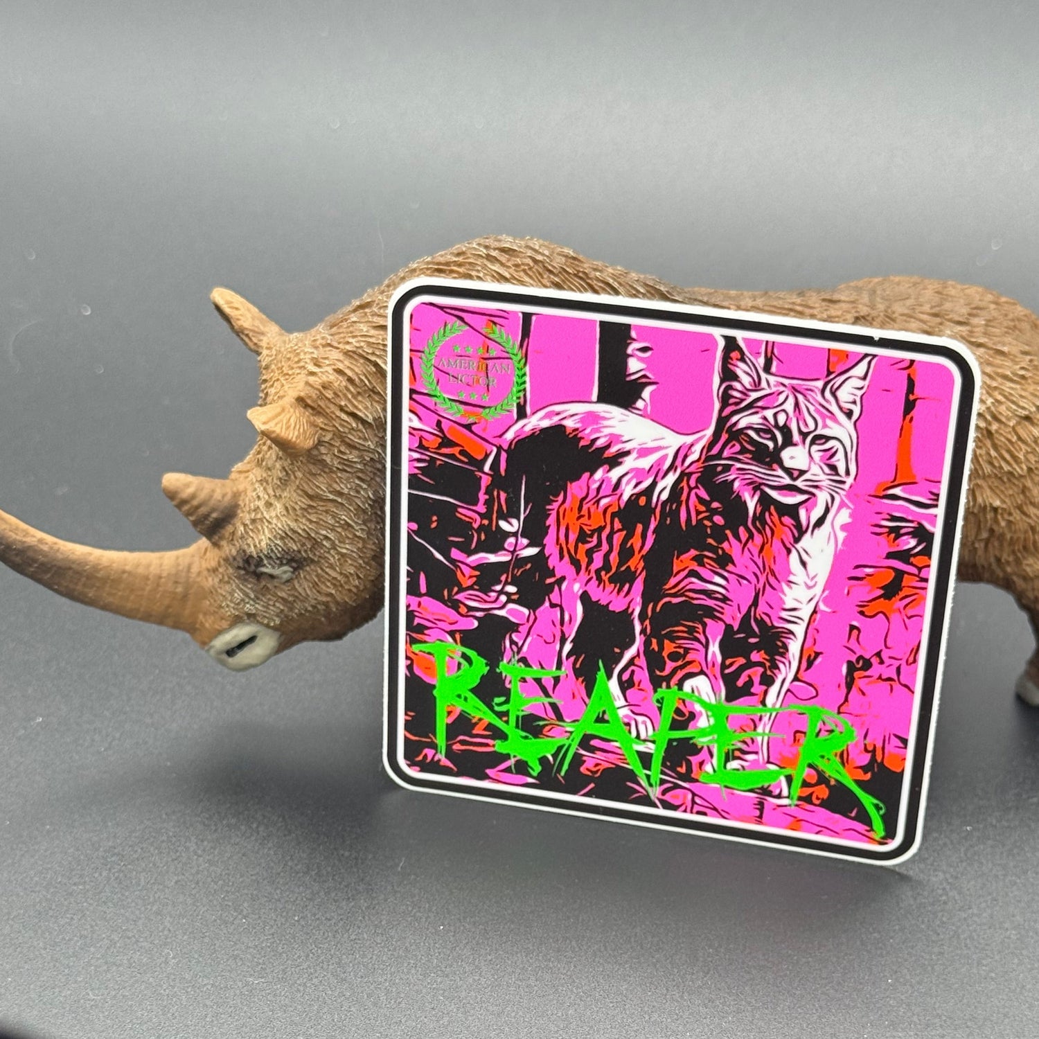 Black, green and pink sticker of a bobcat with the text 'Reaper' posed with a wooly Rhino