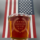 A Shot glass with the American Lictor Logo filled with whiskey in front of an American Flag