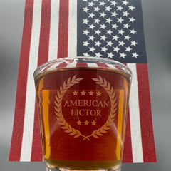 A Shot glass with the American Lictor Logo filled with whiskey in front of an American Flag