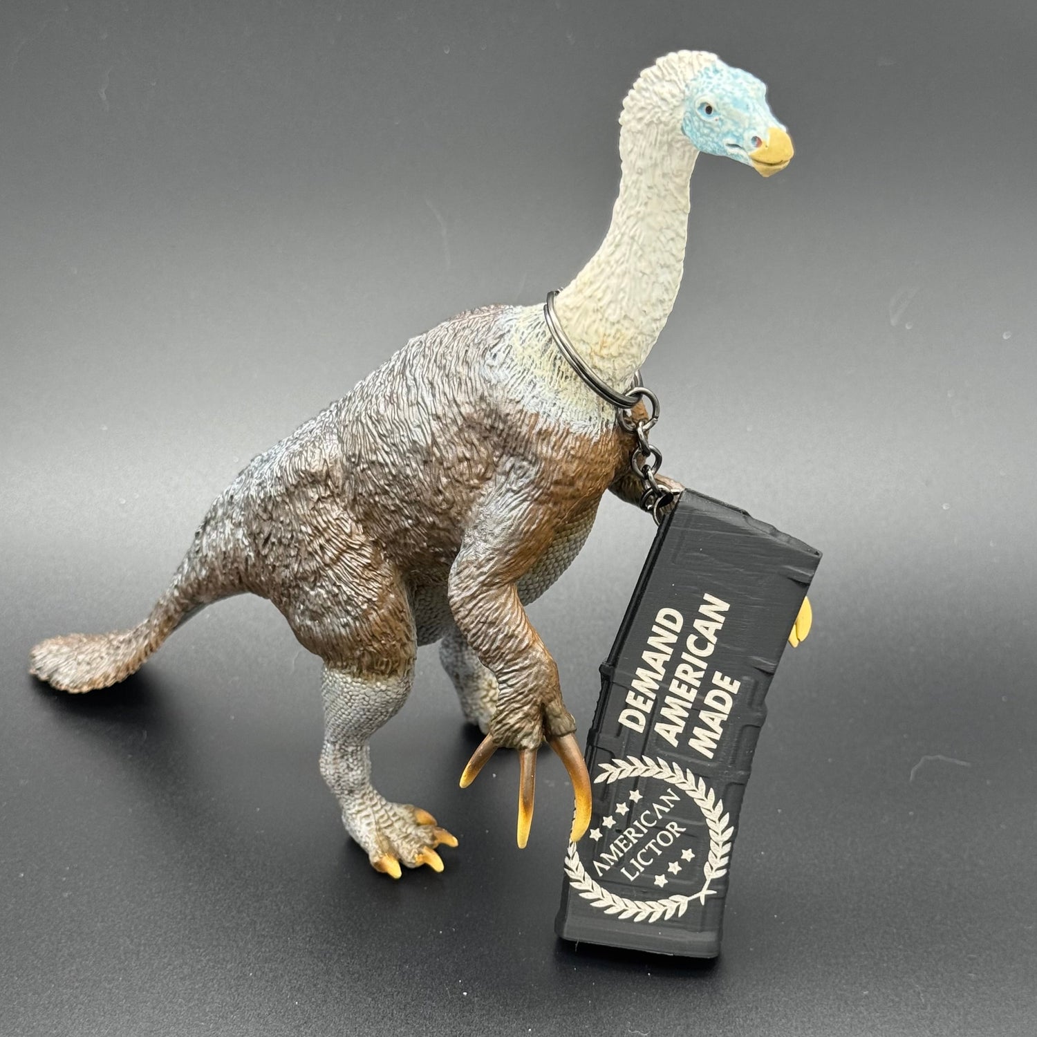 A black keychain shaped like an AR15 mag with the American Lictor logo and slogan Demand American Made' posing with a dinosaur