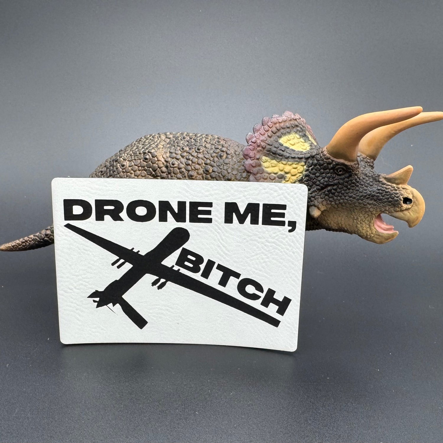 A white leatherette patch of a reaper drone and the text 'Drone Me, Bitch' posed with a dinosaur