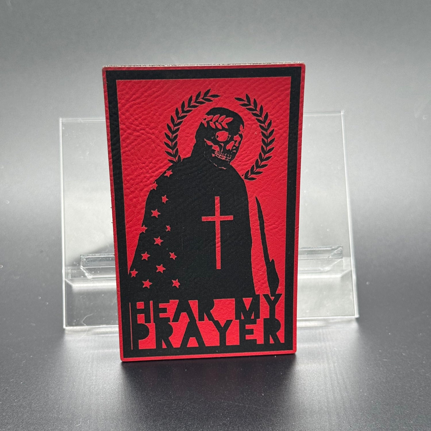 A red leatherette patch with a man with a skeleton mask draped in the American Flag with a Laurel Halo and cross and the text 'Hear my Prayer'