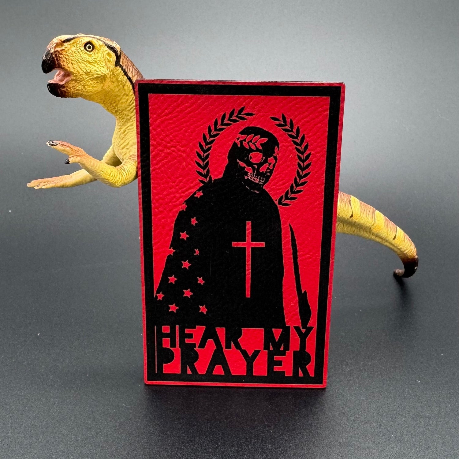 A red leatherette patch with a man with a skeleton mask draped in the American Flag with a Laurel Halo and cross and the text 'Hear my Prayer' posed with a dinosaur
