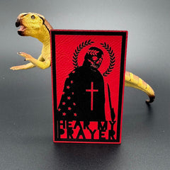 A red leatherette patch with a man with a skeleton mask draped in the American Flag with a Laurel Halo and cross and the text 'Hear my Prayer' posed with a dinosaur