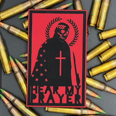 A red leatherette patch with a man with a skeleton mask draped in the American Flag with a Laurel Halo and cross and the text 'Hear my Prayer' on a bed of ammo
