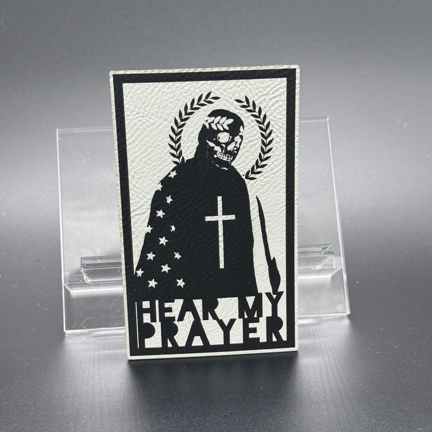 A white leatherette patch with a man with a skeleton mask draped in the American Flag with a Laurel Halo and cross and the text 'Hear my Prayer'