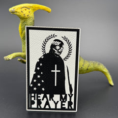 A white leatherette patch with a man with a skeleton mask draped in the American Flag with a Laurel Halo and cross and the text 'Hear my Prayer' posed with a dinosaur