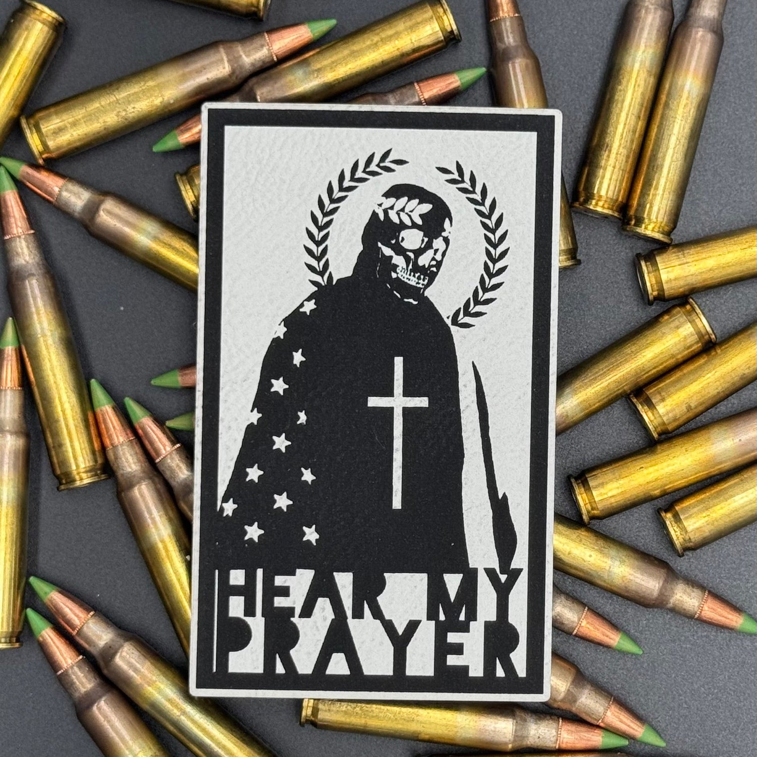 A white leatherette patch with a man with a skeleton mask draped in the American Flag with a Laurel Halo and cross and the text 'Hear my Prayer' on a bed of ammo