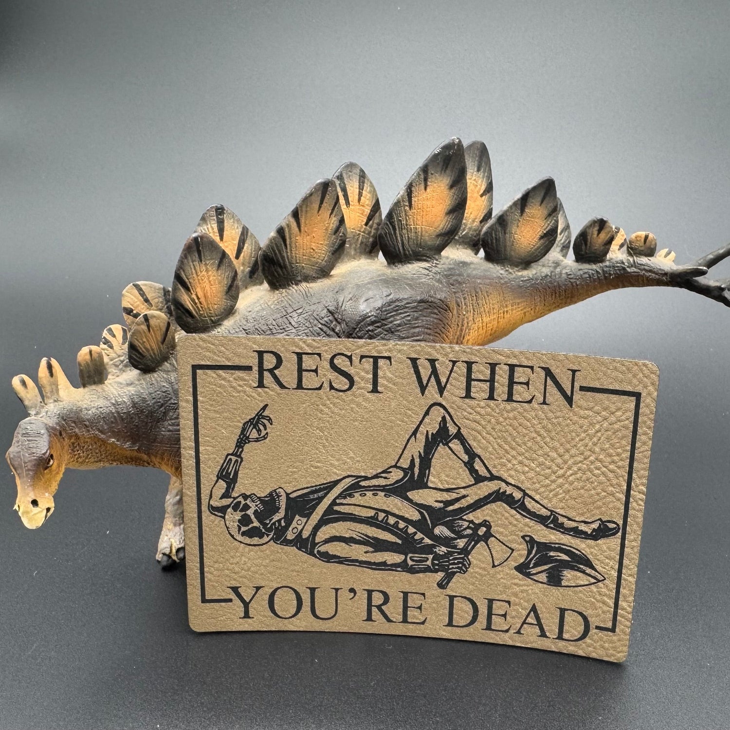 A leatherette patch in beige picturing a skeleton wearing colonial soldier clothing with a tomahawk and the text 'Rest when you're dead