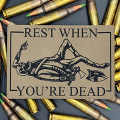 A leatherette patch in beige picturing a skeleton wearing colonial soldier clothing with a tomahawk and the text 'Rest when you're dead" on a bed of ammo