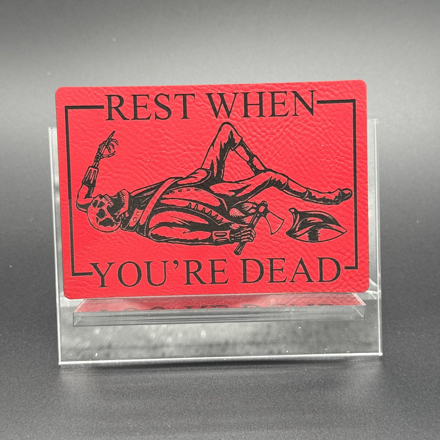 A leatherette patch in red picturing a skeleton wearing colonial soldier clothing with a tomahawk and the text 'Rest when you're dead
