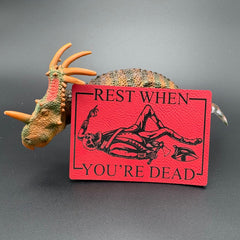 A leatherette patch in red picturing a skeleton wearing colonial soldier clothing with a tomahawk and the text 'Rest when you're dead" posed with a dinosaur