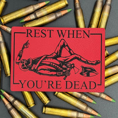 A leatherette patch in red picturing a skeleton wearing colonial soldier clothing with a tomahawk and the text 'Rest when you're dead" on a bed of ammo
