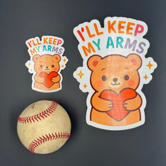 A small and large Sticker of a Brown Cartoon bear holding a heart saying "I'll keep my arms" pictured with a baseball for scale