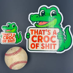 A small and Large Sticker of a Green Cartoon Crocodile holding a sign saying "That's a croc of shit" shown with a baseball for scale