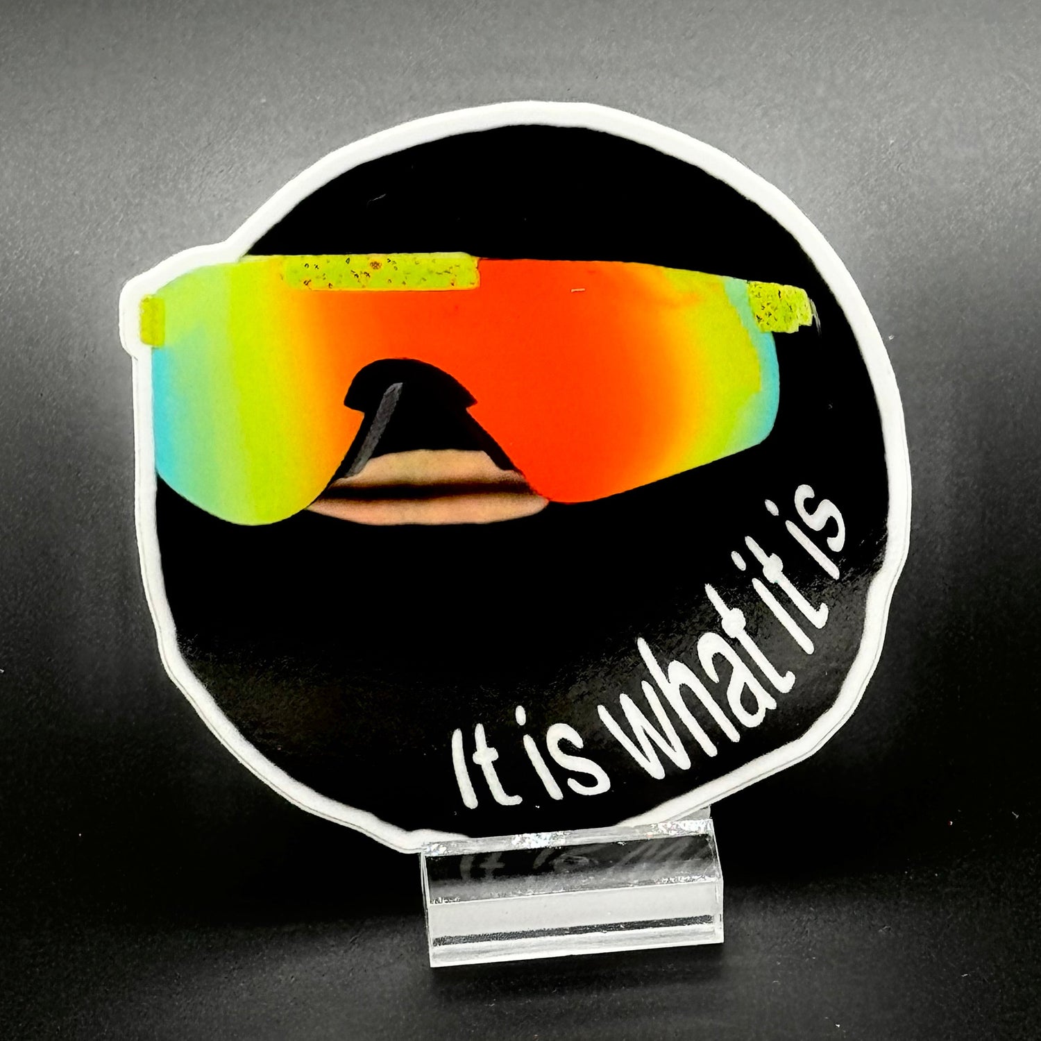 Sticker of a goofy cartoon character wearing reflective sunglasses and wearing a ski mask with the text 'It is what it is'