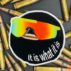 Sticker of a goofy cartoon character wearing reflective sunglasses and wearing a ski mask with the text 'It is what it is' on a bed of ammo