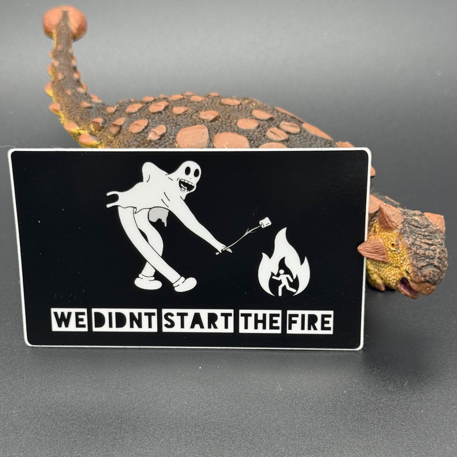 Sticker of a cartoon character in a ghost suit roasting a marshmallow over a fire posing with a dinosaur