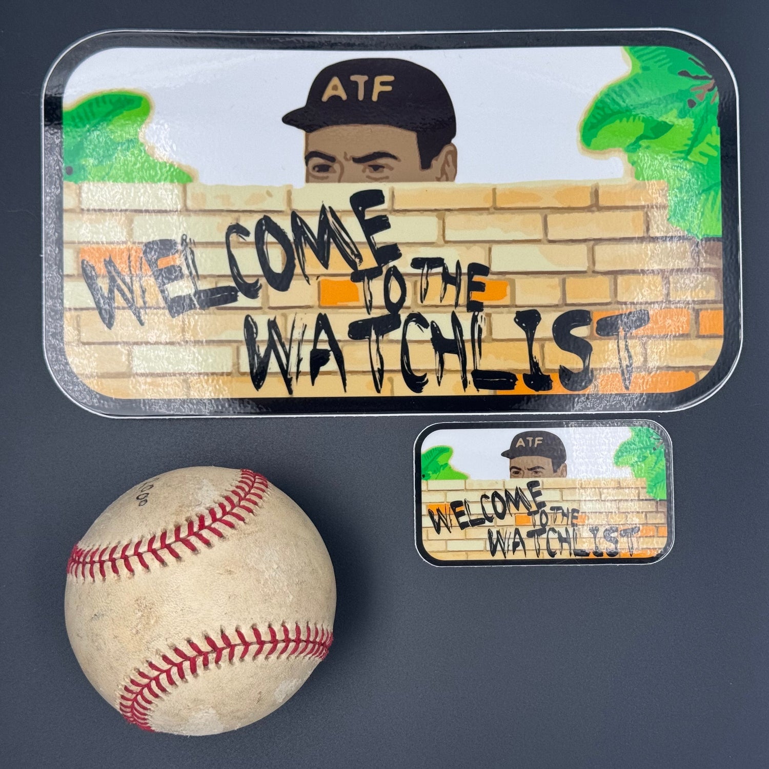 A small and large Sticker of an ATF agent peeking over a wall with the text 'Welcome to the Watchlist' pictured with a baseball for scale