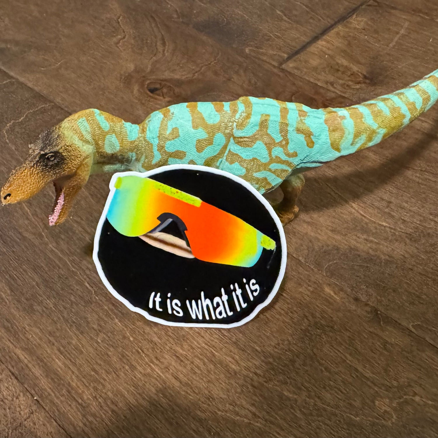 Toy dinosaur next to a sticker with sunglasses and text on a wooden surface