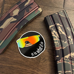 Camouflage magazine on a wooden surface with a sticker featuring sunglasses and text 'It is what it is'