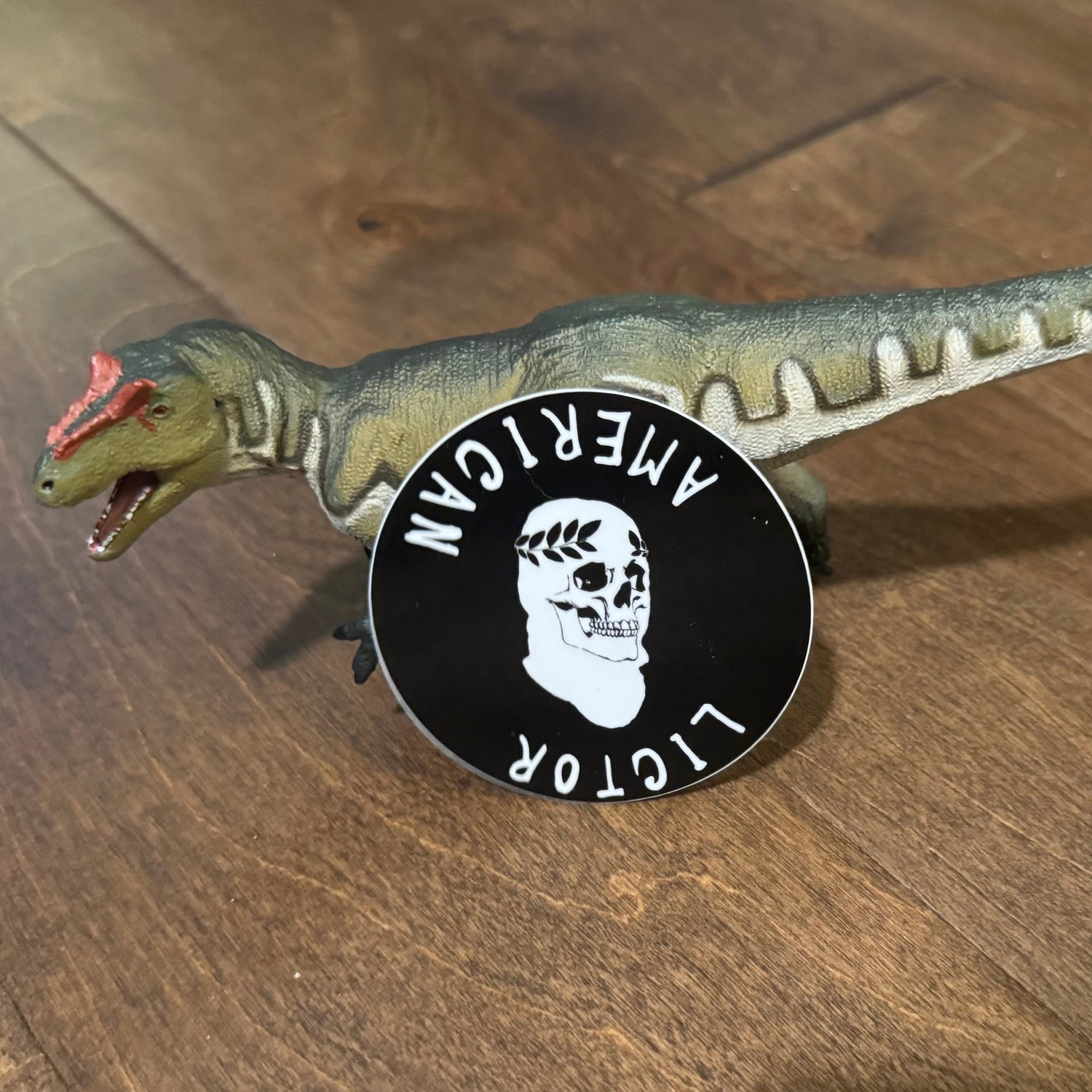 Dinosaur toy with an American Lictor inverted and laurel skull logo on a wooden surface