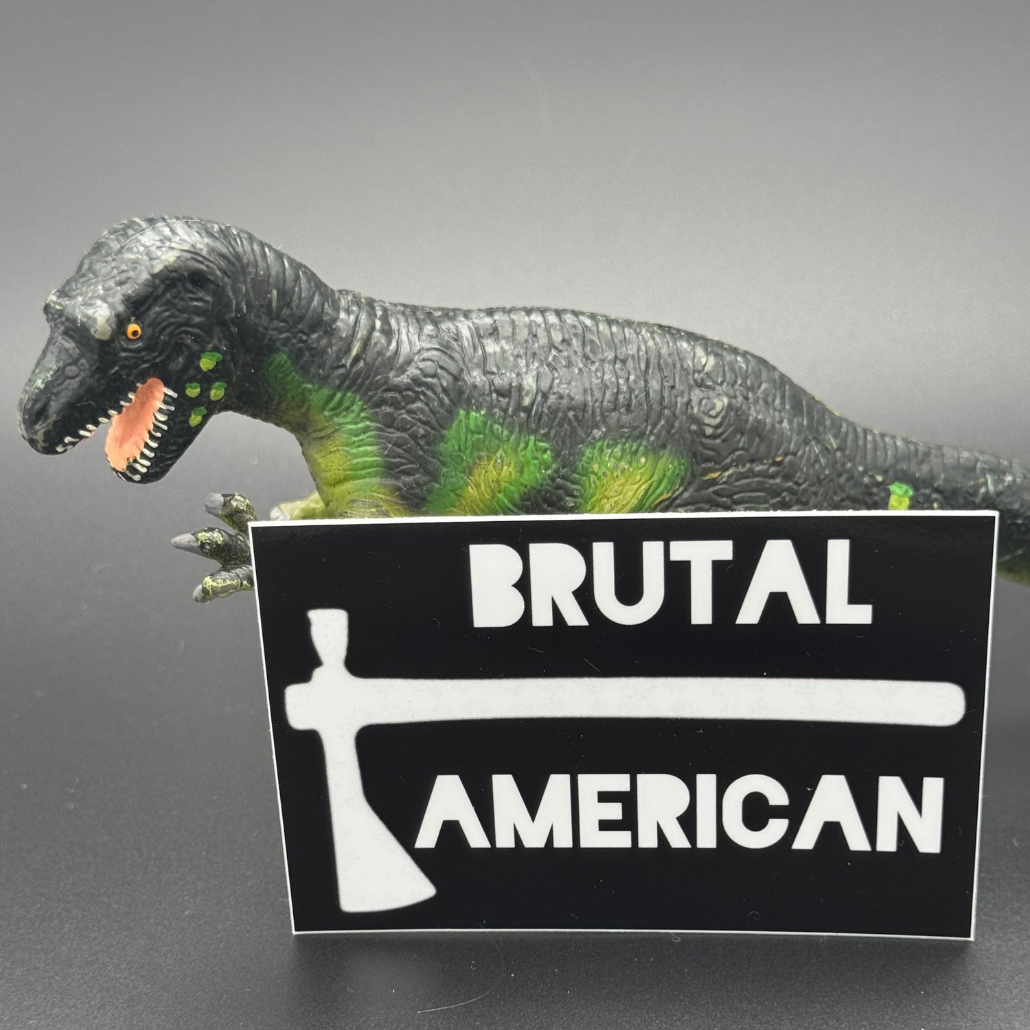 Black sticker with a tomahawk and the text 'Brutal American' posing with a dinosaur