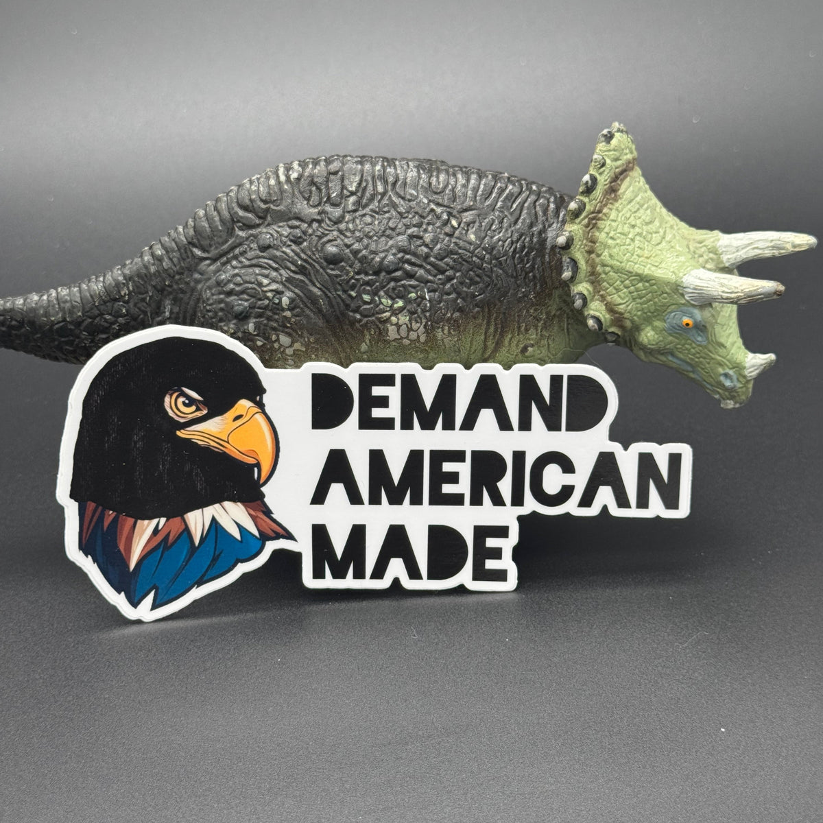 Sticker of an Eagle wearing a ski mask with the text 'Demand American Made' posing with a dinosaur