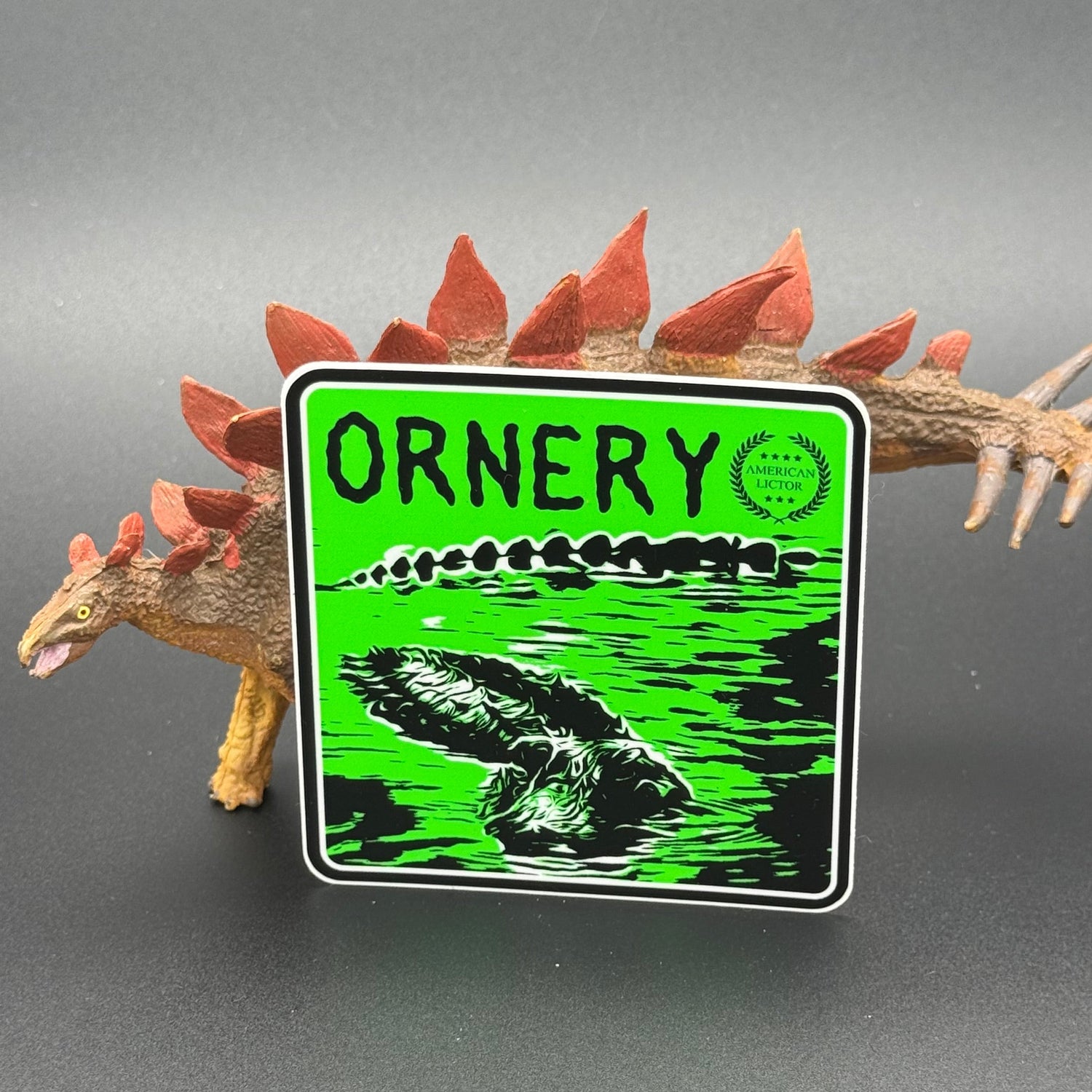 Black and green sticker of an alligator with the text 'Ornery' posed with a dinosaur