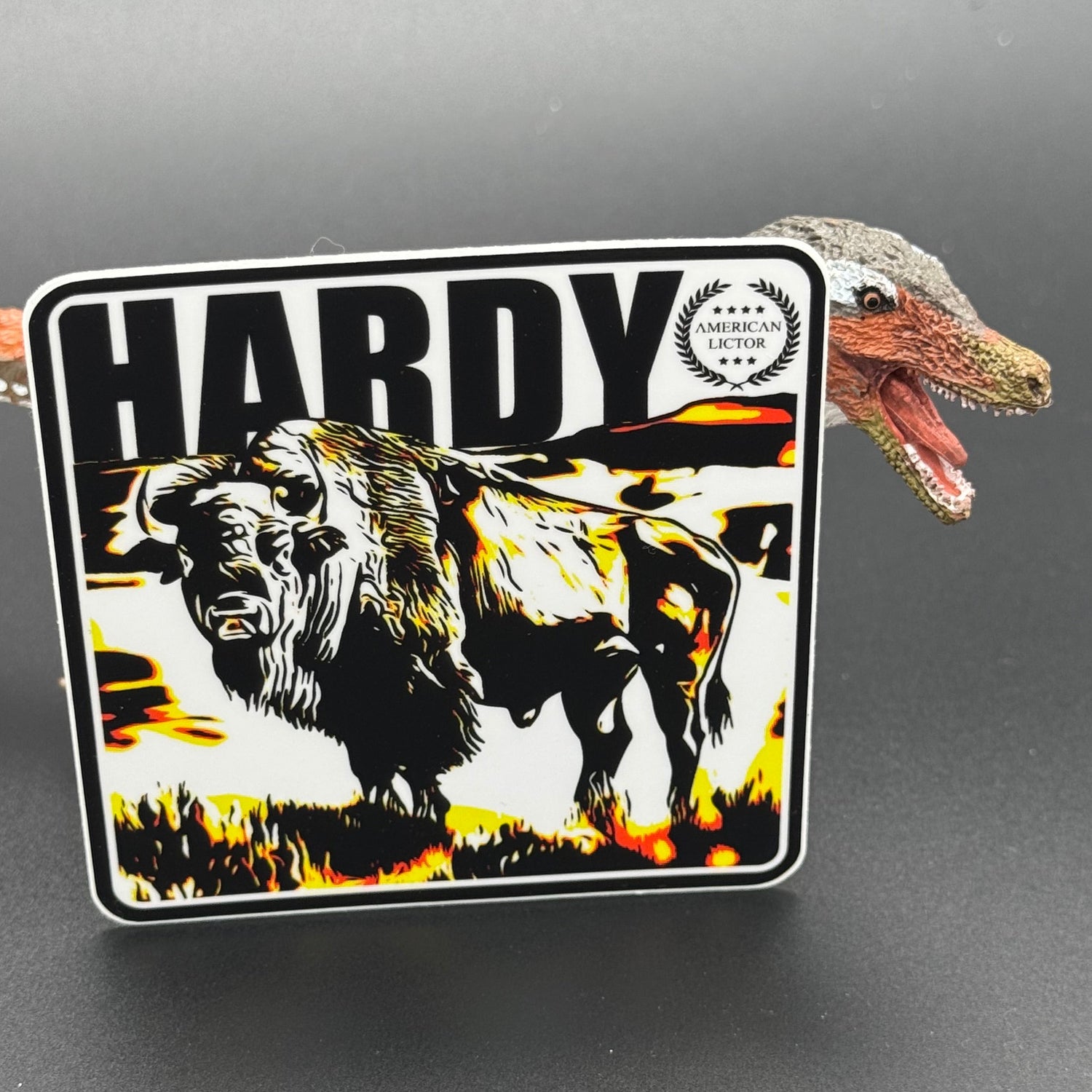 Black, yellow and white sticker of a Bison with the text 'Hardy' posed with a dinosaur