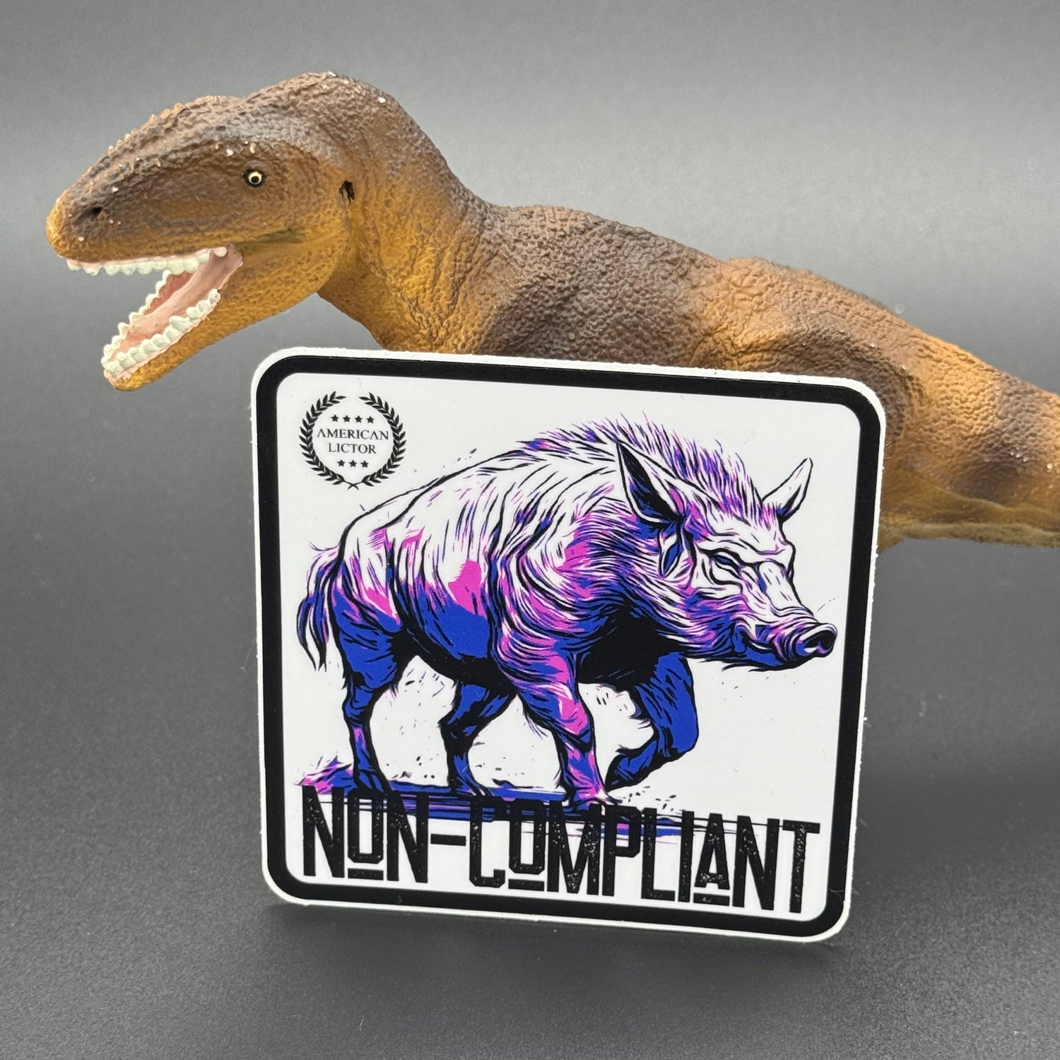 Black, purple, blue and white sticker of a boar with the text 'Non-Compliant' posed with a dinosaur