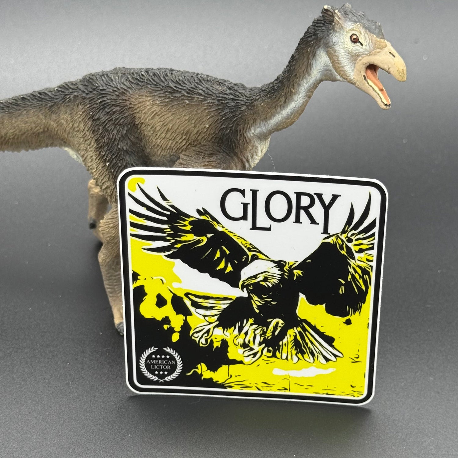 Black, yellow and white sticker of an eagle with the text 'Glory' posed with a dinosaur