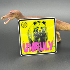 Yellow and pink Sticker of a grizzly bear with the text 'Unruly' posed with a dinosaur