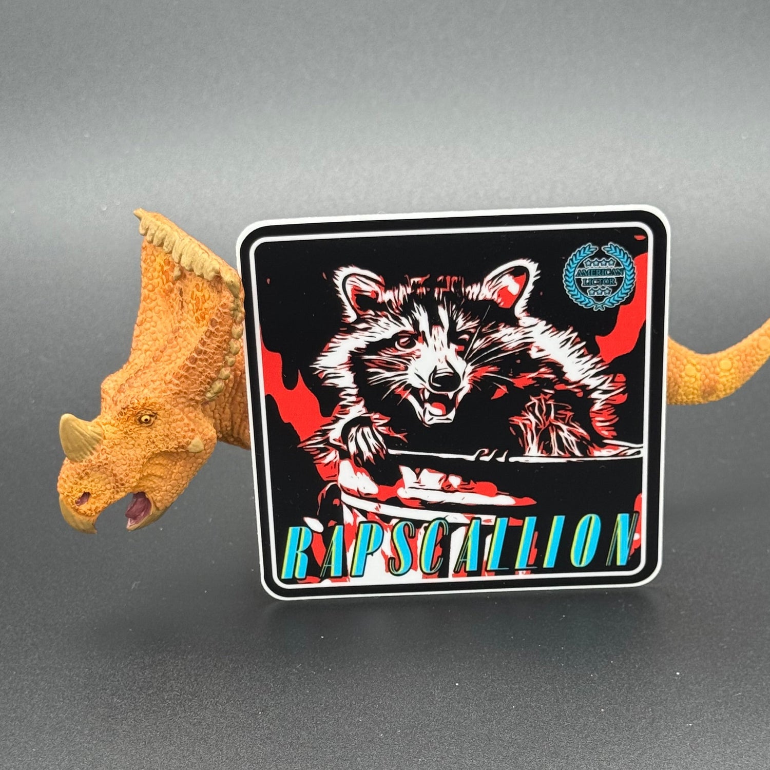Black, red and teal Sticker of a Raccoon with the text 'Rapscallion' posed with a dinosaur